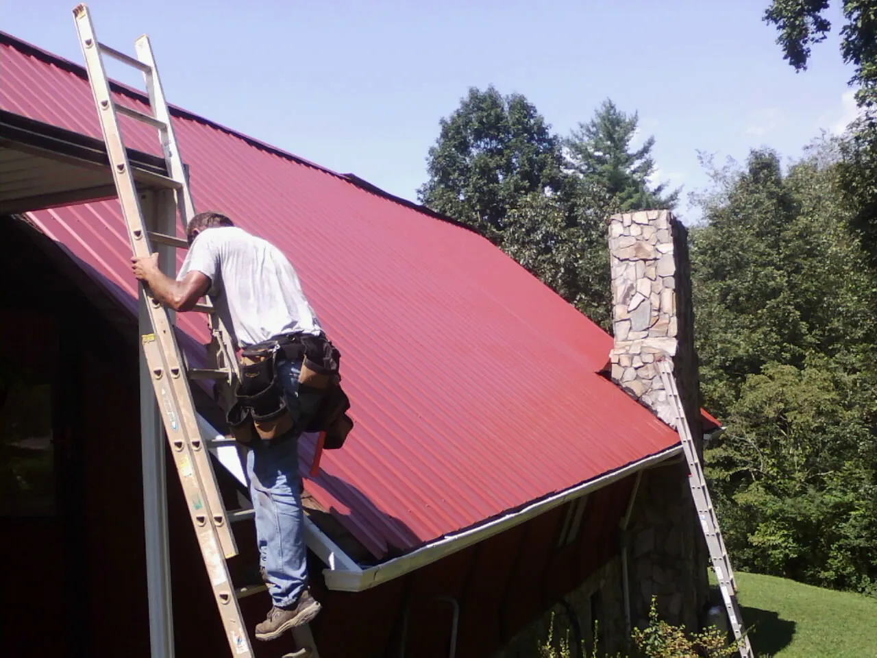 Roofer on a ladder beside a red metal roof and stone chimney — Soffit & Fascia Repair by Roofing Brothers in Halstad